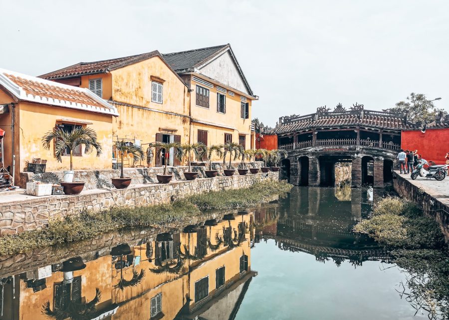 A bright red Japanese Covered Bridge over the river next to a yellow building, Hoi An Ancient Town, Vietnam