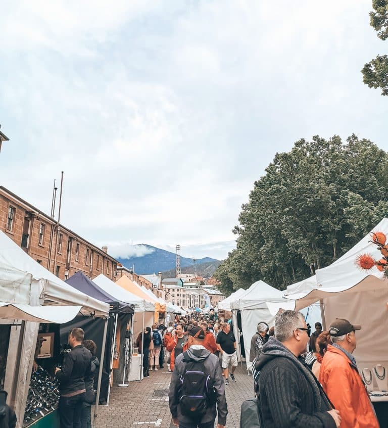 A street of market stalls filled with people browsing and a mountainous backdrop, Salamanca Market, Hobart, Tasmania