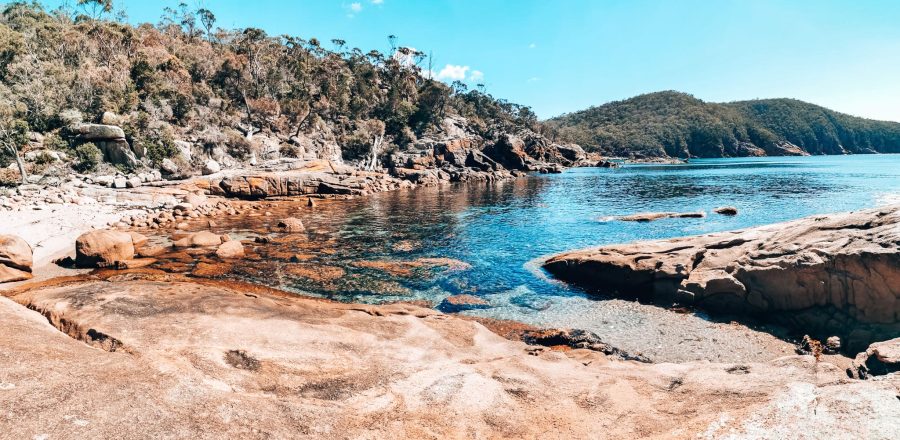 Red rock sloping down into crystal clear ocean with mountains in the background at Sleepy Bay, Freycinet National Park, best places to visit in Tasmania