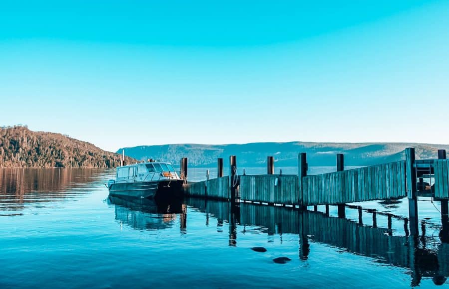 A boat next to a jetty on the calm waters of Lake St Clair surrounded by greenery, Tasmania, Australia