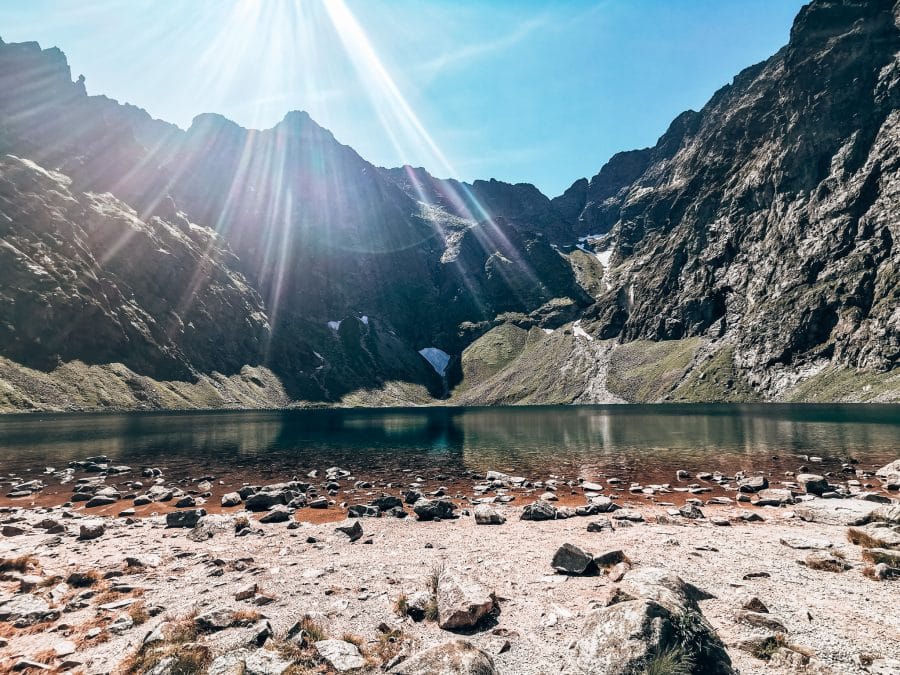 A lake nestled underneath jagged mountain peaks with the sun shining above, Czarny Staw, Hiking Rysy Mountain, Poland