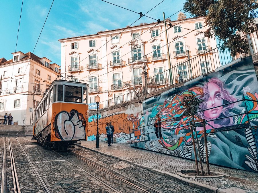 A steep cobbled street with a yellow tram and vibrant street art, must see in Lisbon Portugal