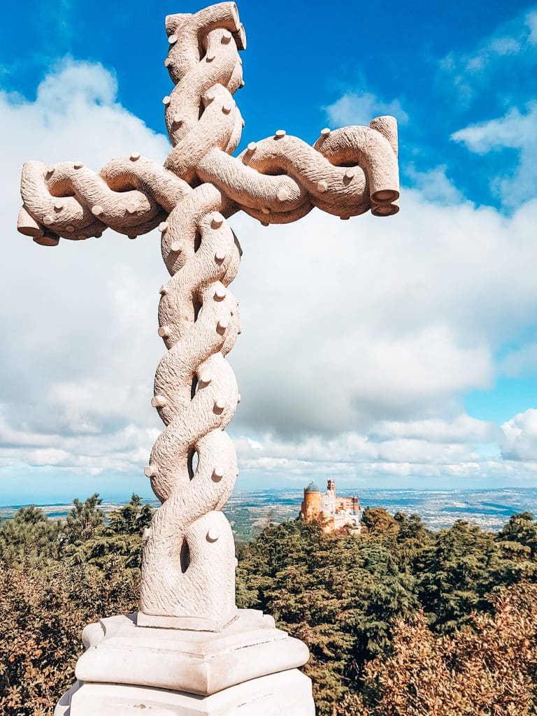 A tall cross and the colourful Pena Palace protruding through the forest at Cruz Alta in Sintra, Places you must see in Lisbon, Portugal