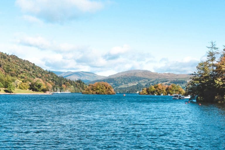 The calm waters of Lake Windermere with mountains looming in the distance and golden autumn colours on the trees, Bowness, Lake District, England, UK