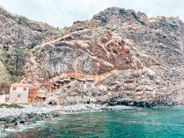 The view from the deck out across the pristine water, pebbly beach and magnificent ancient cave houses carved into the side of a gigantic volcanic cliff, Calhau da Lapa, Madeira, Portugal