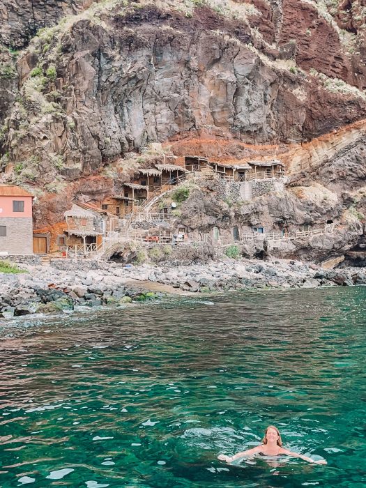 Helen swimming in the sea in front of traditional fishermen's cave houses built into the side of a cliff at Calhau da Lapa, best hidden gems in Madeira, Portugal