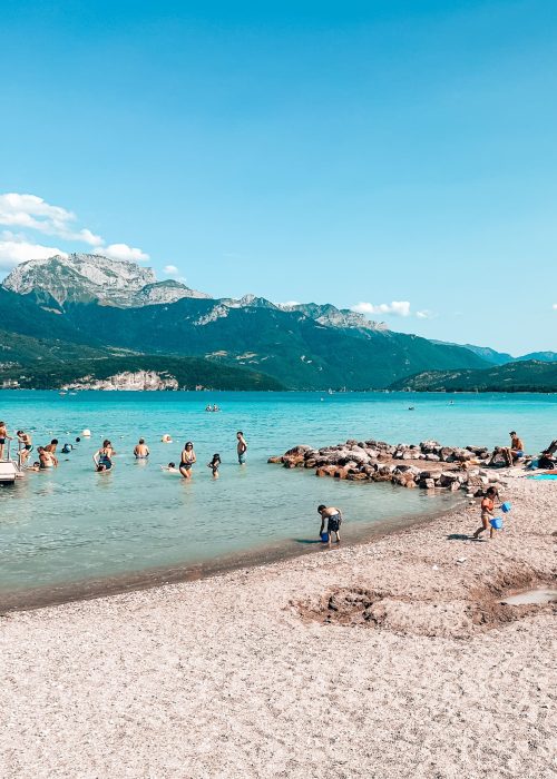 The piercing blue water of Lake Annecy with a mountainous background and lots of people swimming or on the beach, Annecy, France