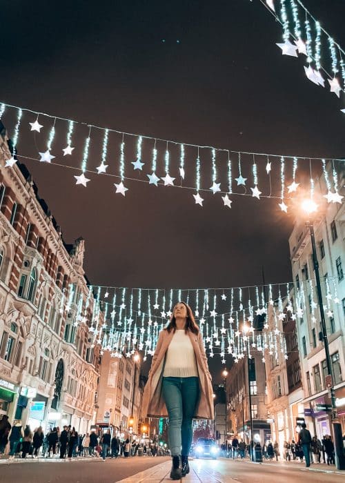 Helen walking in the middle of the road on Oxford Street with thousands of twinkly stars strung between the buildings above her making these some of the top Christmas lights in London