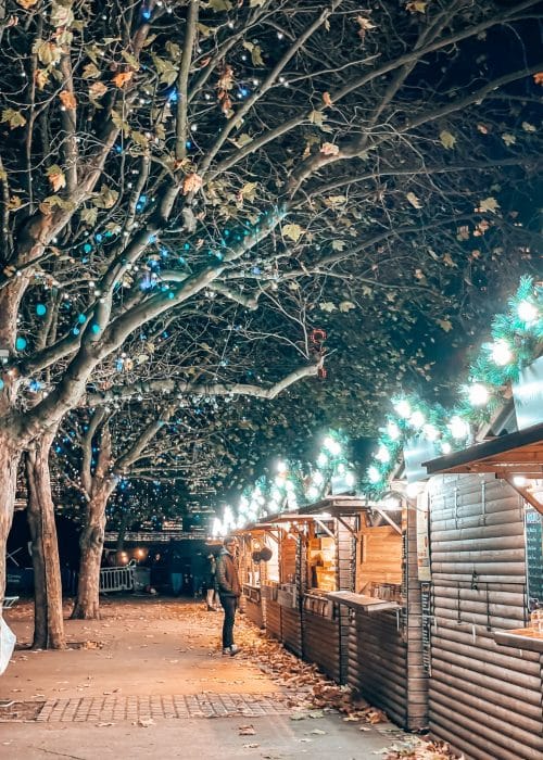 A row of wooden cabins with sparkling lights on the roofs and twinkly lights in the trees at Southbank Centre Winter Market in London