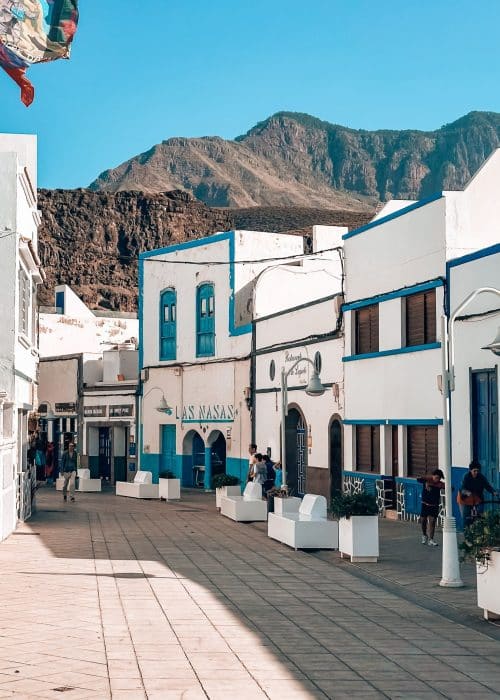A row of white and blue buildings on a narrow street with dramatic mountain peaks as a backdrop, Puerto de las Nieves