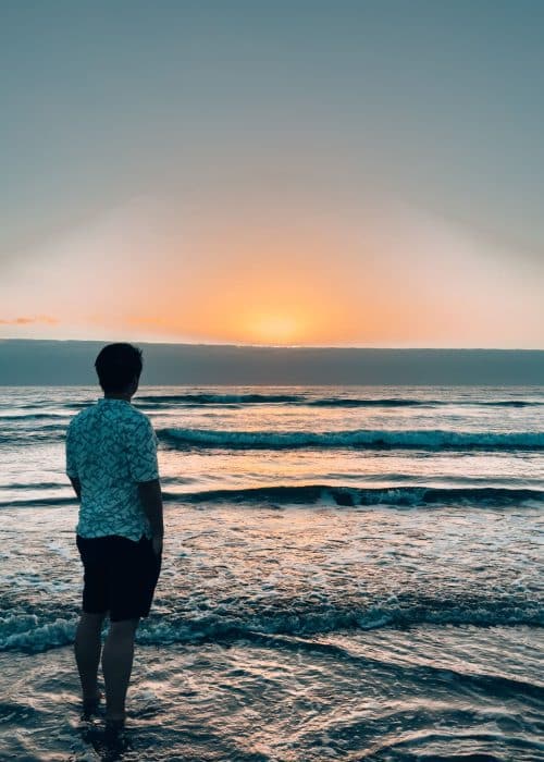 Andy stood at the edge of the sea with waves washing over his feet at sunrise at the Dunas de Maspalomas, Gran Canaria, Canary Islands, Spain