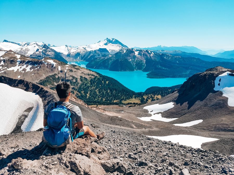 Andy taking a break on volcanic scree looking far below at the turquoise Garibaldi Lake and snow-capped mountains, Whistler