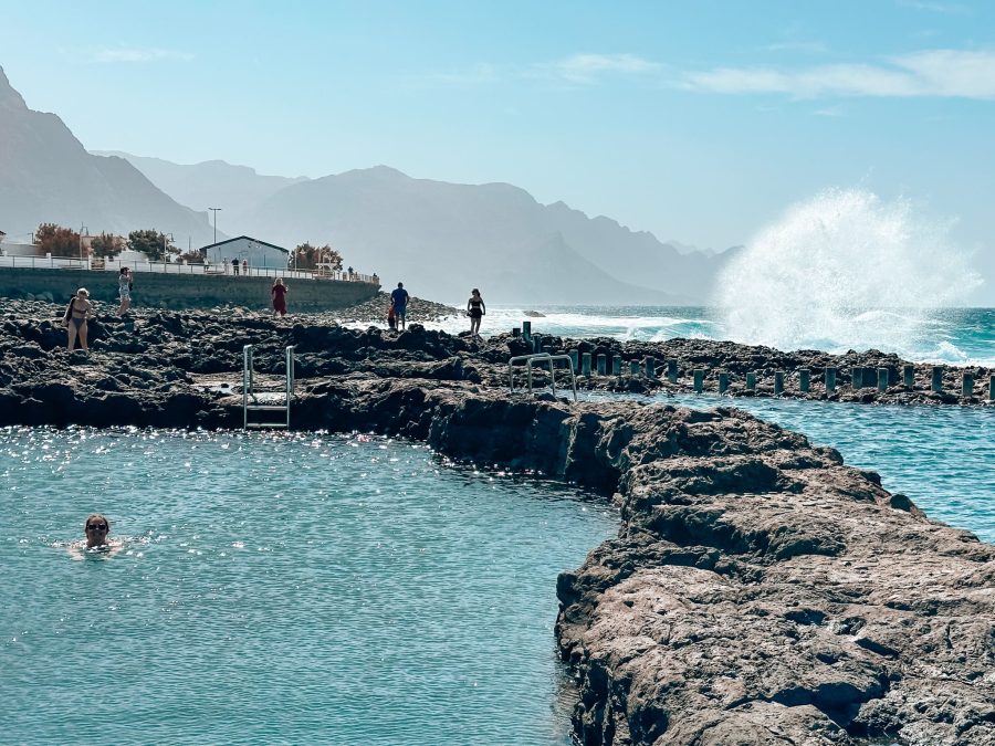 Helen swimming in Las Salinas de Agaete with huge waves crashing into the rocks behind and rugged cliffs plunging into the ocean, Puerto de las Nieves, Gran Canaria