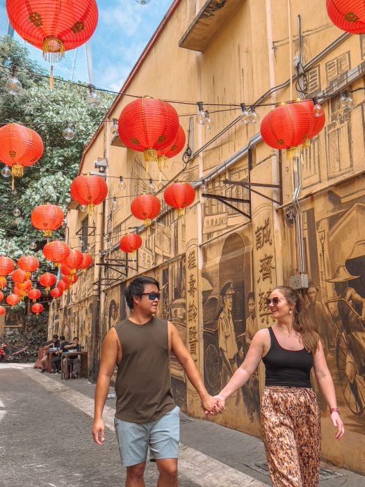 Helen and Andy walking hand-in-hand down a narrow road with street art with red lanterns hanging between the buildings, Chinatown, Kuala Lumpur