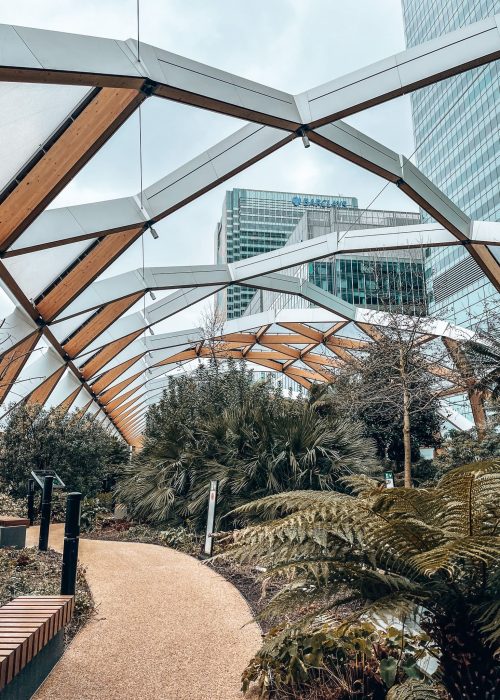 A glass roof with skyscrapers towering above it and plants inside, Crossrail Place Roof Garden, London Itinerary