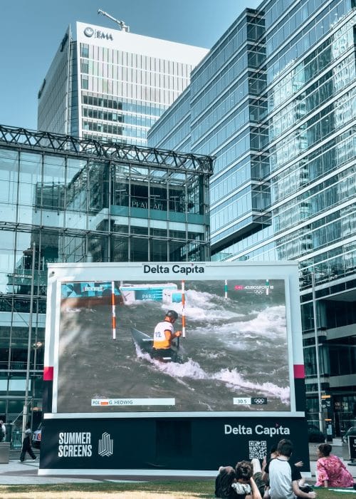 An outdoor TV screen in front of glass skyscrapers at Canada Square Park, Canary Wharf, London