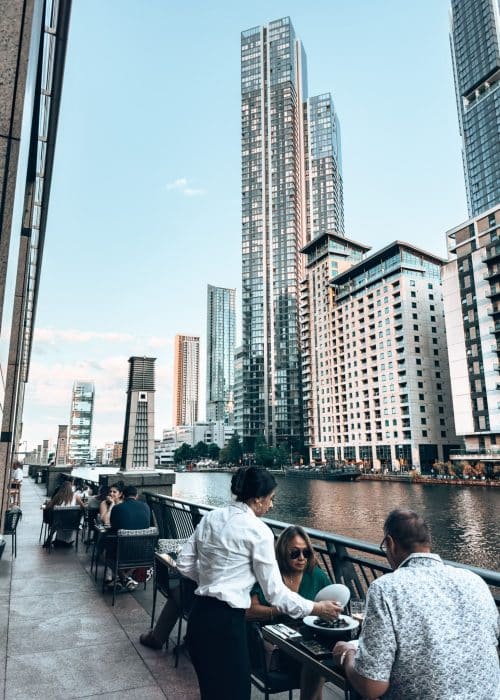 A restaurant's outside terrace next to the water framed with huge glass skyscrapers, Canary Wharf, London Itinerary