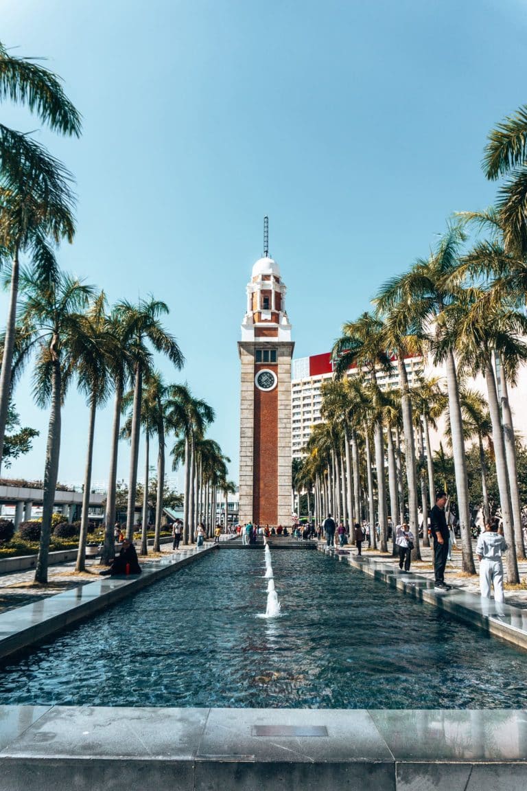 A red granite clock tower at the end of a water feature lined with palm trees, The Former Kowloon-Canton Railway Clock Tower, Hong Kong