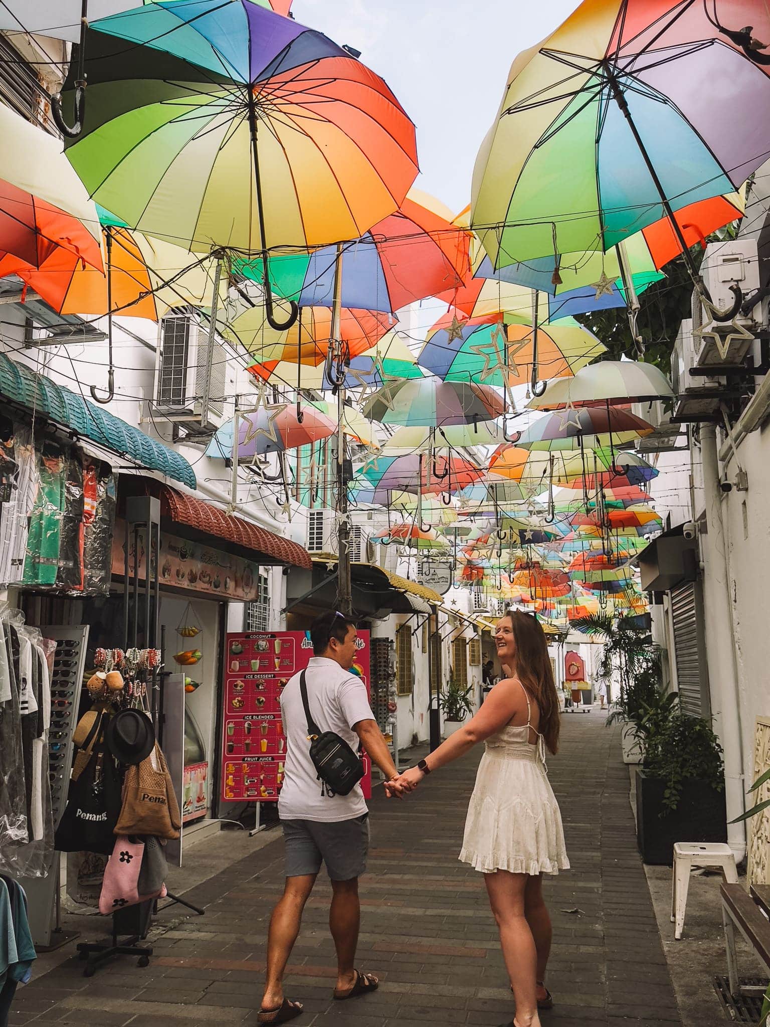 Helen and Andy holding hands with colourful umbrellas hanging above them on a narrow street, Umbrella Alley, Places to Visit in Penang, Malaysia